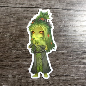 Op de afbeelding: Een groen en bruin cartoonfiguur met gloeiende ogen en een kroon van bladeren. Het figuur draagt een donkergroene robe en houdt een groen blad in zijn hand.