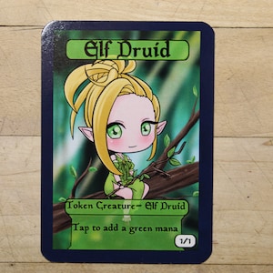 May include: A green and blue Magic: The Gathering card with an illustration of a cartoon elf druid with blonde hair and green eyes. The card text reads "Elf Druid", "Token Creature - Elf Druid", "Tap to add a green mana", and "1/1".