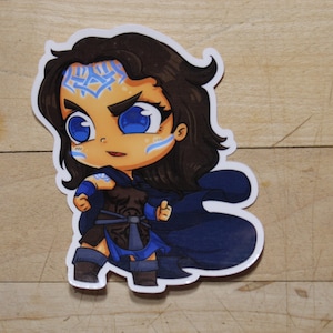 Puede incluir: Una pegatina de dibujos animados azul y marrón de una mujer con el pelo largo y oscuro, que lleva una capa azul y un atuendo azul y marrón. Tiene los ojos azules y un diseño azul y blanco en la frente.