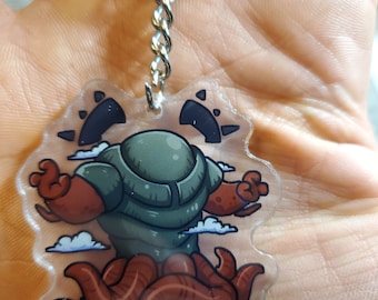 May include: A clear acrylic keychain featuring a cartoon illustration of a green and brown creature with red arms and tentacles. The creature has a large head with two black eyes and a white cloud behind it.
