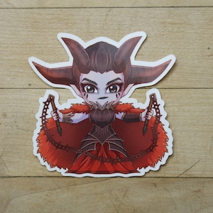 May include: A sticker featuring a cartoon illustration of a female demon with red horns, a red cape, and black armour. She is holding two spiked chains in her hands.