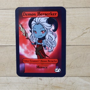 May include: A trading card depicting a cartoon demon berserker with blue skin, white hair, and horns. The card has a red background with the text "Demon Berserker" and "Menace". The card is 2/3.