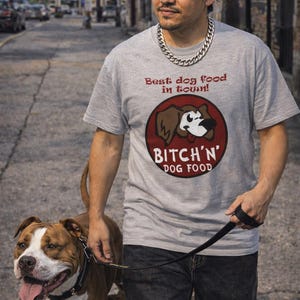 May include: A light gray t-shirt with the text "Best dog food in town!" and a graphic of a dog's face inside a red circle. The person is wearing a silver chain, sunglasses, and a bandana. A brown and white dog is on a leash.