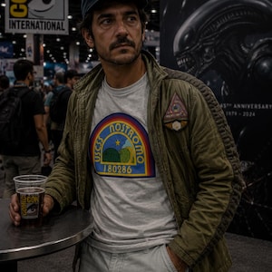 May include: A navy blue baseball cap with a circular patch, a light gray t-shirt featuring a colorful USCSS Nostromo 180286 graphic, a triangular patch on the chest, and an olive green utility jacket with quilted shoulder detailing and light gray cargo pants.