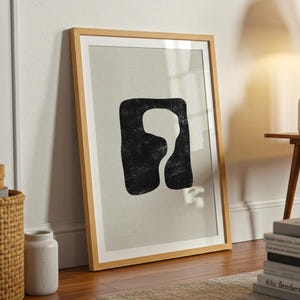 Puede incluir: Una lámina de arte enmarcada con fondo gris claro y una forma abstracta negra irregular en el centro. La obra está dentro de un marco de madera clara y presenta un aspecto texturizado y pintado a mano dentro de la forma negra.