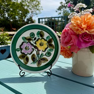 May include: A ceramic plate with a floral pattern in purple, yellow, and green, set on a black metal stand. The plate has a green border. A vase of colourful flowers is in the background. The scene is set outdoors on a blue table.