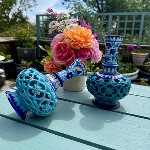 May include: Two ornate ceramic vases, one turquoise and one blue and white, with floral designs. The turquoise vase has openwork. A bouquet of pink and orange flowers sits nearby. The vases are on a light blue table.