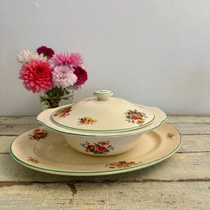 May include: A cream-coloured vintage serving dish with lid, placed on a matching plate. The dish and plate have a floral pattern with red, yellow, and orange flowers. A green trim borders the edges. A vase of colourful flowers is in the background.