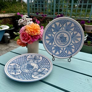 May include: Two white ceramic plates with blue floral patterns. One plate is displayed on a black metal stand. A bouquet of orange and pink roses sits in a white vase on a light blue table. The plates have a decorative, hand-painted appearance.