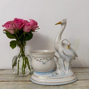 May include: A white ceramic swan planter with a light blue base and gold trim. The planter has a cup-shaped holder and is decorated with blue floral accents. A glass vase with pink roses is to the left.