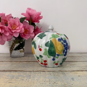 May include: A white ceramic lidded jar with a ribbed, pumpkin-like shape, decorated with a hand-painted pattern of yellow pears, green leaves, blue grapes, and small red berries, topped with a small, pointed finial on the lid.