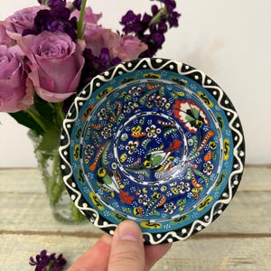 May include: A small round ceramic bowl featuring a vibrant blue background with intricate hand-painted floral and bird motifs in red, yellow, green, and white. The rim is decorated with a black and white scalloped pattern, creating a bold contrast.