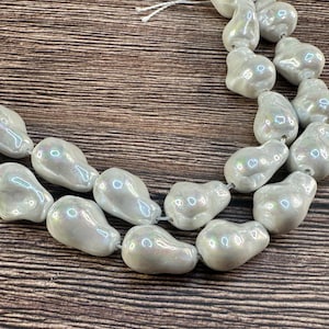 May include: Strand of iridescent, pear-shaped beads. The beads are a light, pearly color with a subtle sheen, strung together on a thin, white cord. The beads are arranged on a brown, wood-grain surface.
