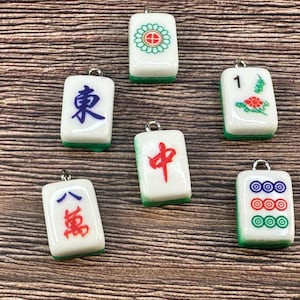 Può includere: Sei ciondoli rettangolari a forma di tessera del Mahjong con faccia bianca e retro verde, ciascuno dotato di un anellino metallico sulla parte superiore. Le tessere presentano vari simboli, tra cui un fiore rosso, caratteri blu e cerchi.