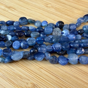 May include: A strand of polished, irregular, pebble-shaped blue kyanite beads. The beads exhibit varying shades of deep navy, royal blue, and light sky blue, with a smooth, glossy finish and natural internal inclusions visible throughout the stones.