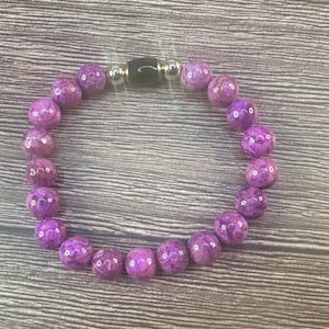 May include: A purple beaded bracelet with a black and silver clasp. The bracelet features round, polished beads in varying shades of purple. The beads are strung together to form a complete circle. The bracelet is displayed on a wooden surface.
