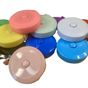 Puede incluir: Una colección de cintas métricas retráctiles redondas y coloridas en varios tonos, incluyendo rosa, verde, amarillo, azul y naranja. Cada cinta tiene un botón central y una pequeña pestaña. Útiles para manualidades y costura.
