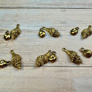 May include: Gold-colored seashell charms and small round charms are scattered on a light wood surface. The charms have a detailed, textured design. These charms are suitable for jewelry making or craft projects.