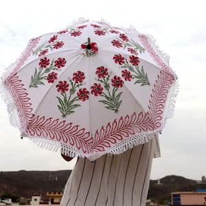 May include: A white parasol with a red and green floral print and white fringe. The parasol is held by a person wearing a striped shirt.