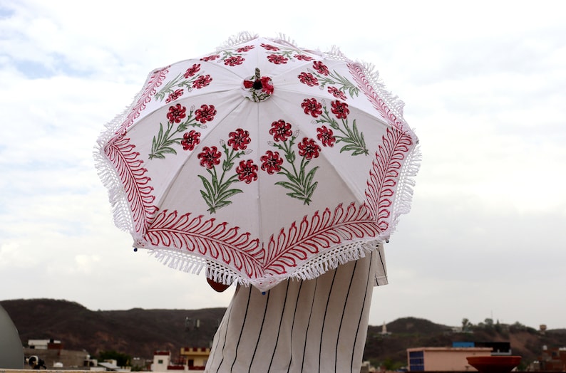 May include: A white parasol with a red and green floral print and white fringe. The parasol is held by a person wearing a striped shirt.