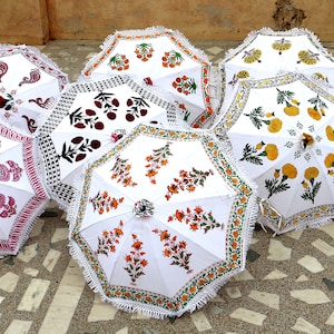 May include: Nine white cotton umbrellas with colorful floral patterns. The umbrellas have white fringe trim and are arranged in a row on a tiled floor.