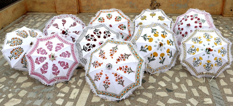May include: Nine white cotton umbrellas with colorful floral patterns. The umbrellas have white fringe trim and are arranged in a row on a tiled floor.