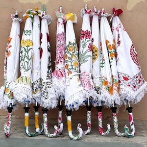 May include: A collection of eight white umbrellas with colorful floral patterns and white fringe. The umbrellas are hanging on a wall.