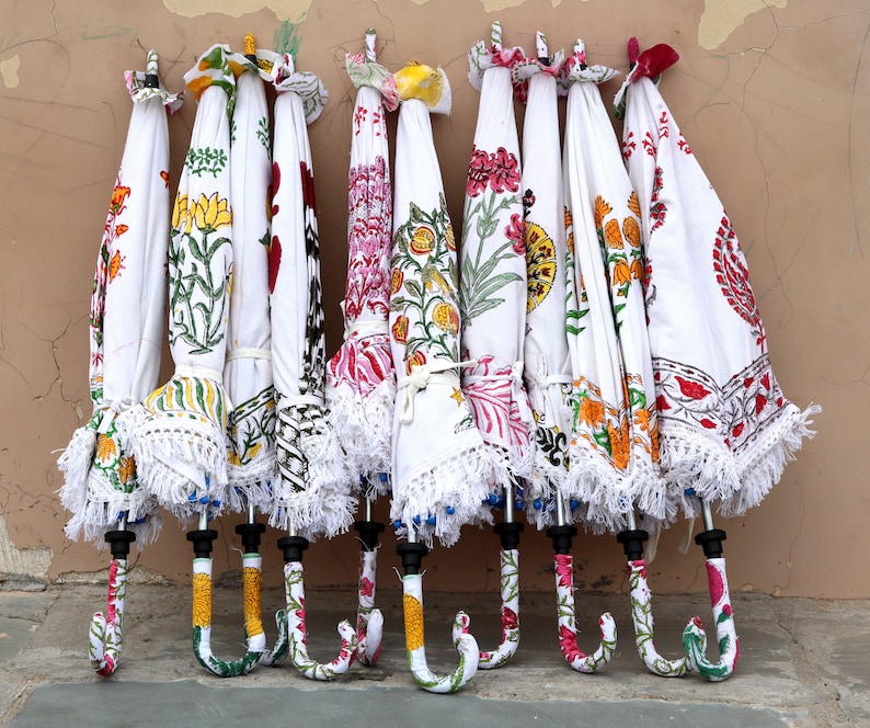 May include: A collection of eight white umbrellas with colorful floral patterns and white fringe. The umbrellas are hanging on a wall.