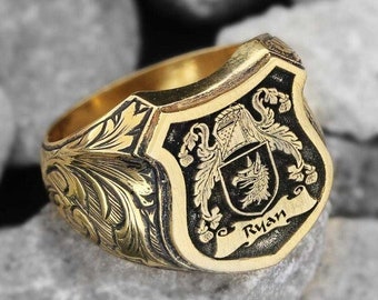 May include: A gold ring with a black shield design featuring a crest and the name "Ryan". The ring is decorated with intricate floral engravings.