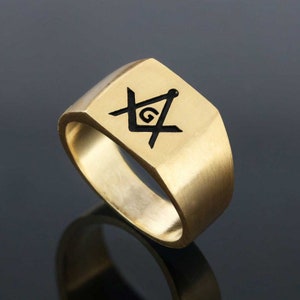 May include: A gold ring with a black square and compass design, featuring a 'G' in the center.