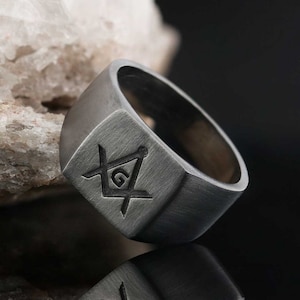 May include: A silver ring with a square face featuring a Masonic symbol engraved in the center. The ring has a brushed finish.