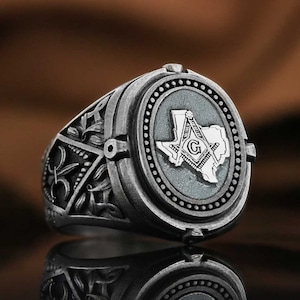 May include: A silver ring with an ornate design featuring a compass and square, a 'G' and the outline of the state of Texas. The ring has a dark gray finish.