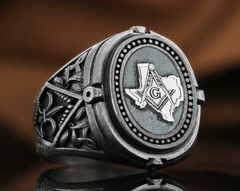 May include: A silver ring with an ornate design featuring a compass and square, a 'G' and the outline of the state of Texas. The ring has a dark gray finish.