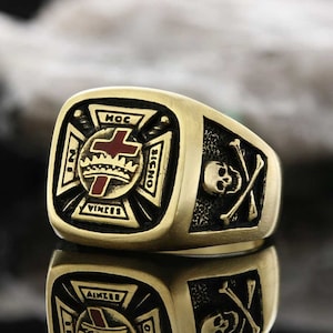 Könnte beinhalten: Ein goldener Ring mit einer quadratischen Oberfläche, die ein rotes Kreuz mit einer Krone und den Worten "HOC SIGNO VINCES" zeigt. Die andere Seite des Rings hat einen Totenkopf und gekreuzte Knochen.