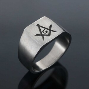 May include: A silver ring with a square face featuring a black engraved Masonic symbol with a 'G' in the center.