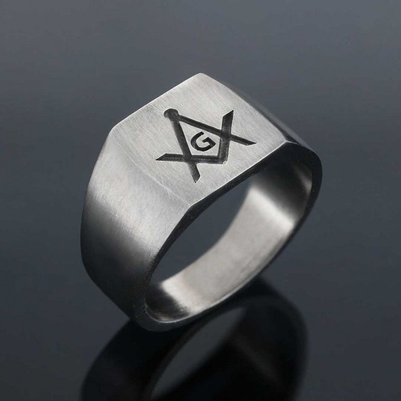 May include: A silver ring with a square face featuring a black engraved Masonic symbol with a 'G' in the center.