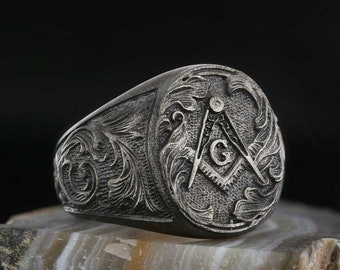Può includere: Un anello d'argento con un simbolo massonico inciso e dettagli floreali intricati. L'anello è di forma ovale e ha una finitura testurizzata.