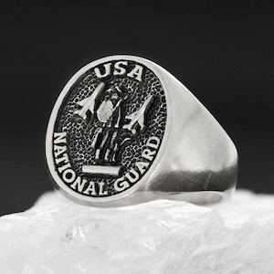 May include: A silver ring with a round, textured top featuring the words "USA NATIONAL GUARD" and a stylized image of a military aircraft.