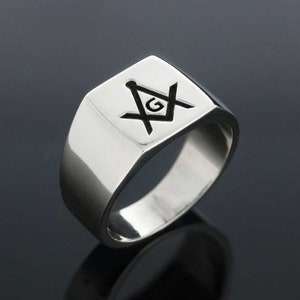 May include: A silver ring with a square face featuring a black Masonic symbol with a 'G' in the center.