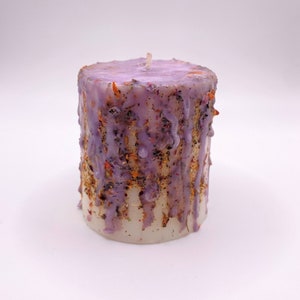 Puede incluir: Una vela cilíndrica con base blanca y parte superior lavanda. La vela tiene un diseño de cera goteada con motas doradas y naranjas. Una sola mecha está centrada en la parte superior de la vela. La vela está sobre un fondo blanco.