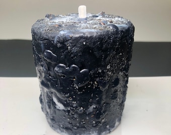 May include: A textured black candle with a white wick, resting on a white rectangular tray. The candle has a rough, uneven surface with white and silver accents. The candle is cylindrical and appears handmade.