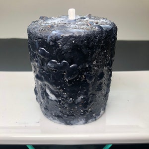 May include: A textured black candle with a white wick, on a white rectangular tray. The candle has a rough, uneven surface with white and silver accents. The candle is cylindrical and appears handmade.