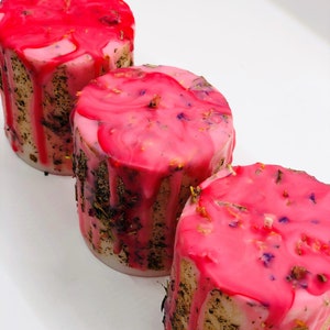 May include: Three cylindrical candles with a pink, dripped wax top. The candles have a light-colored base with dried herbs and flowers embedded in the sides. The pink wax is decorated with dried flowers.