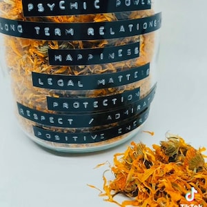 以下が含まれることがあります： オレンジ色の乾燥したカレンデュラの花びらで満たされたガラス瓶。瓶には、「カレンデュラ」、「サイキックパワー」、「長期的な関係」、「幸福」、「法的問題」、「保護」、「尊敬」、「賞賛」、「ポジティブエネルギー」というラベルが貼られています。
