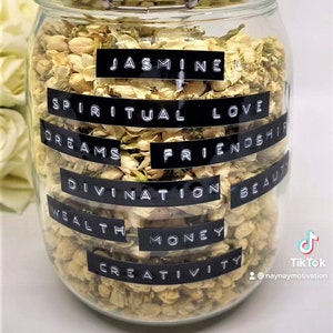 Puede incluir: Un tarro de cristal transparente lleno de flores de jazmín secas. Etiquetas negras con texto blanco enumeran palabras como "Jasmine", "Spiritual Love" y "Dreams". El tarro tiene un cierre metálico y una tapa de cristal.