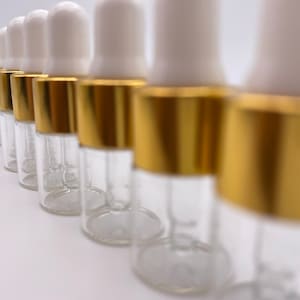 May include: A row of clear glass bottles with gold lids and white droppers. The bottles are lined up in a row on a white surface.