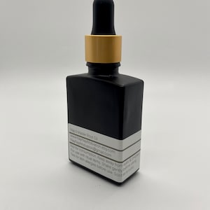 May include: A black square glass bottle with a dropper, gold collar, and black top. The bottle has a white label with text: "Nay's Master Root Oil". The bottle is on a white background.