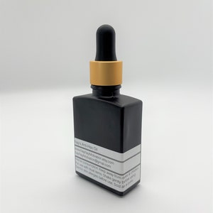 May include: A black square glass bottle with a dropper top. The dropper has a black rubber bulb and a gold-colored metal collar. A white label with black text is affixed to the bottle. The bottle is filled with a liquid.