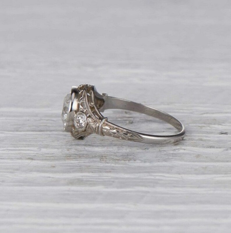 May include: A vintage-style engagement ring with a large, oval-shaped center stone and two smaller accent stones. The ring is made of silver and has intricate details.