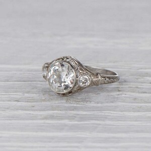May include: A vintage engagement ring with a large, round, clear center stone and two smaller, round, clear accent stones. The ring is set in a delicate filigree design.